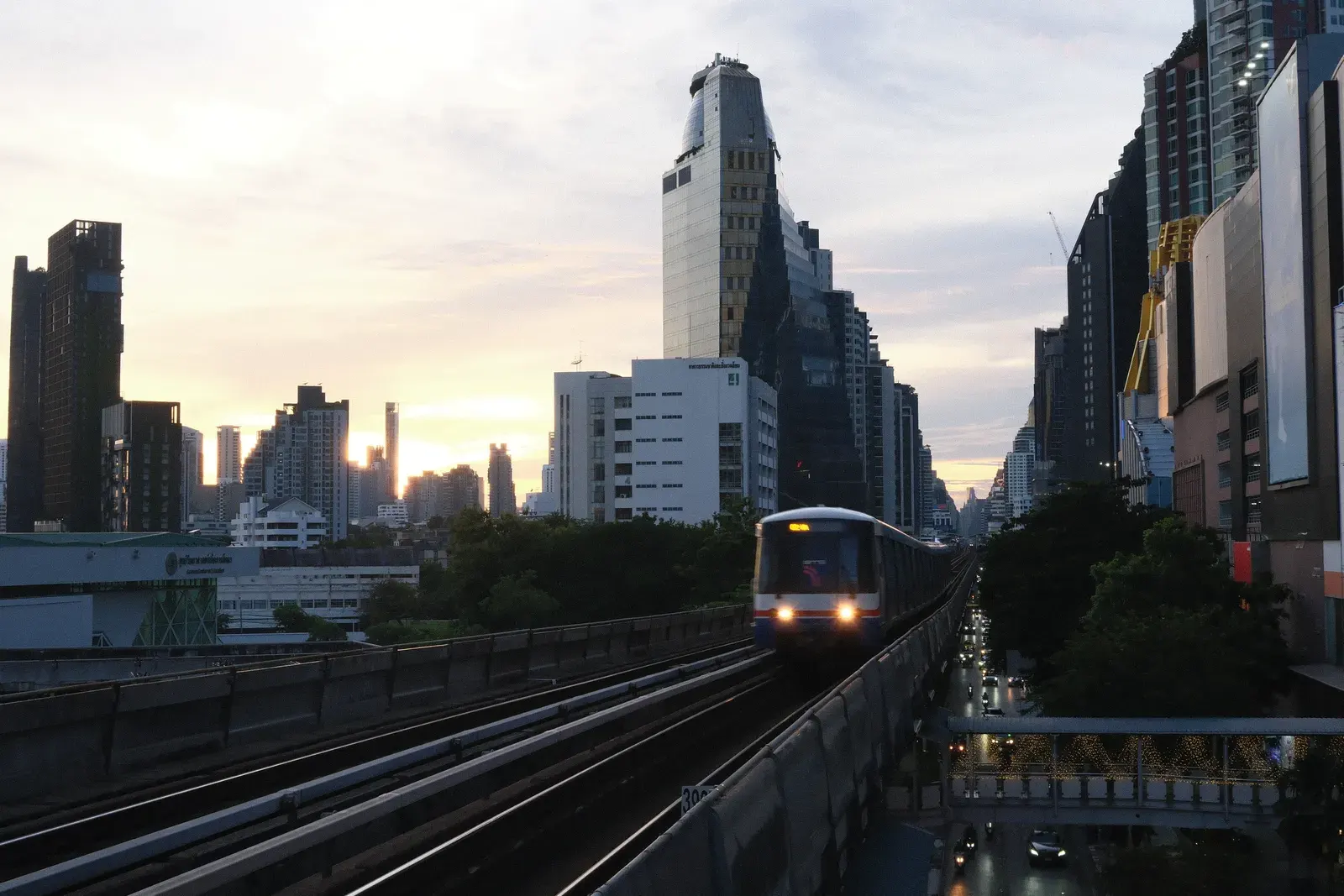 A BTS Skytrain on elevated tracks against the Bangkok skyline during sunset.