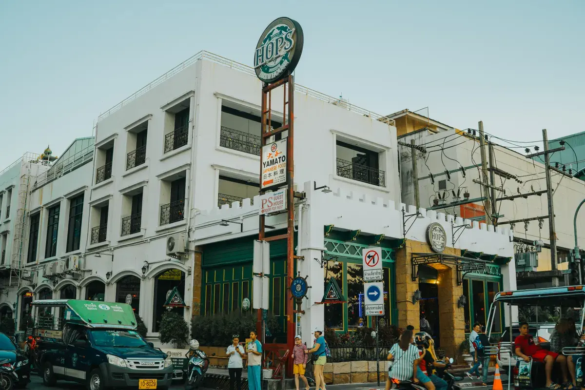 Lively urban street in Bangkok with vehicles and pedestrians near a white building at sunset.