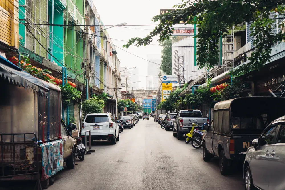 Bustling street in Bangkok with colorful buildings and parked vehicles on a sunny day.