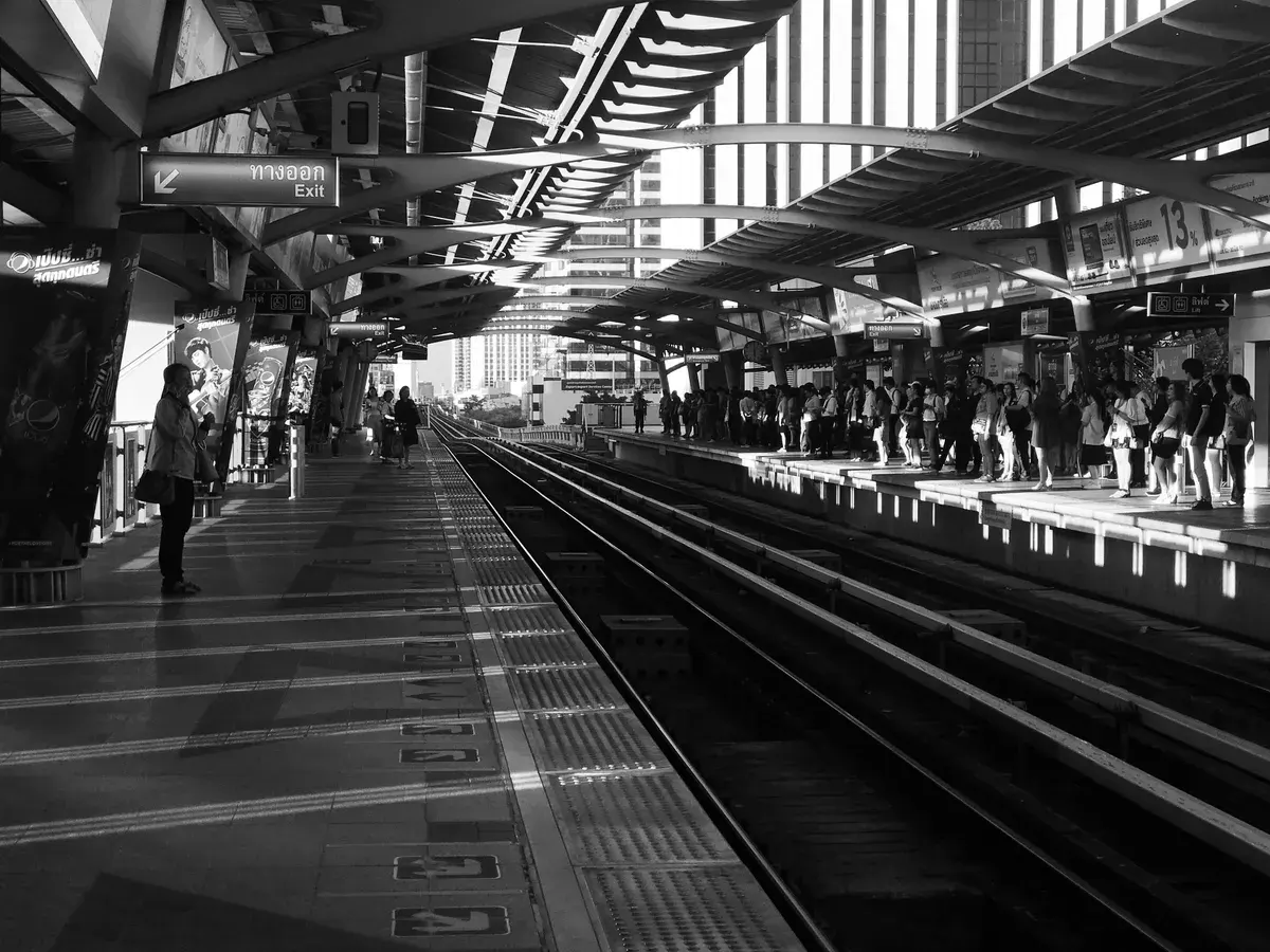 Commuters waiting on a Bangkok train station platform in grayscale lighting.