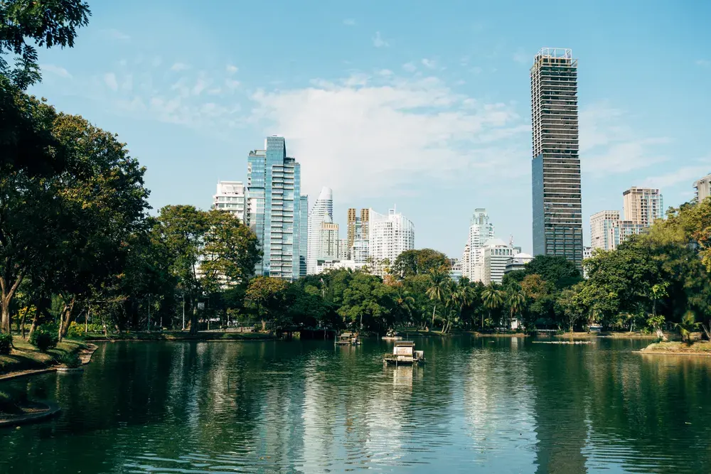 Cityscape view of Bangkok's skyline reflecting in a peaceful park lake under a bright blue sky.