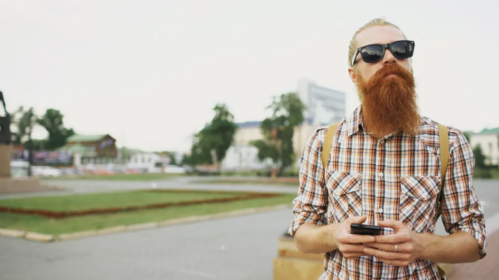 Casual man with a long beard and sunglasses using a smartphone outdoors in an urban setting.