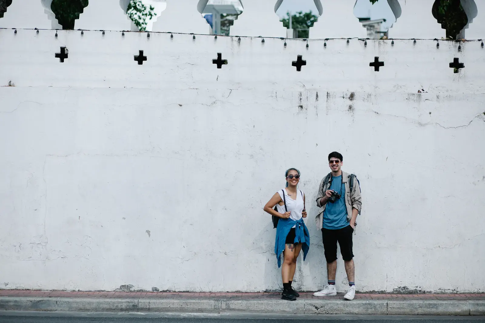 A happy couple in casual wear posing against a historic white wall during their travels.