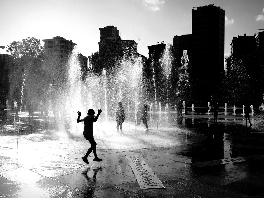 Silhouettes play in water fountains against İstanbul's urban skyline. Captured in monochrome.