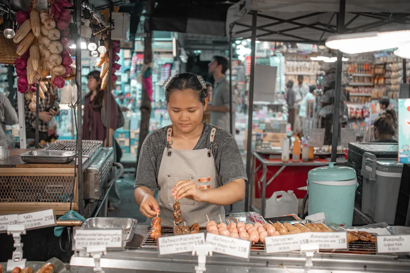 Woman vendor grilling skewers at a bustling Bangkok street market, surrounded by local produce and goods.