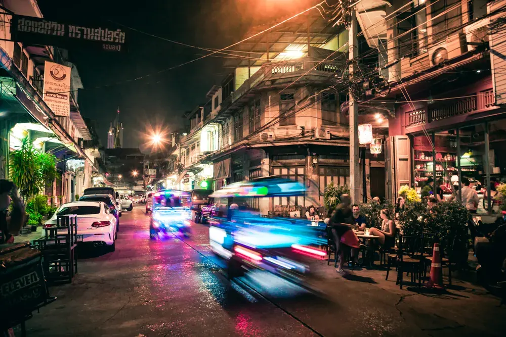 Capture of a lively city street at night with blurred motion from passing tuk-tuks and diners enjoying an outdoor restaurant.
